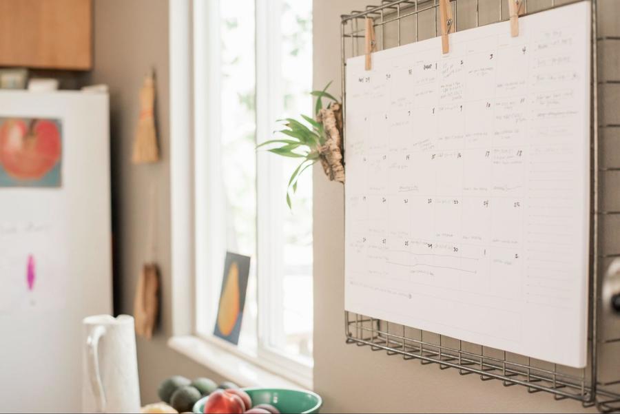Landscape orientated white wall calendar hanging in a kitchen over a wire metal rack affixed with three brown clothespins.