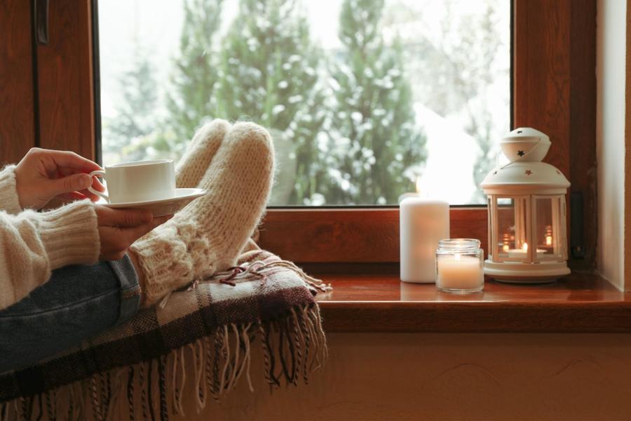 Person holding a white mug while having their white-socked feet up on a wooden windowsill full of candles, looking out to a snowy pined landscape.