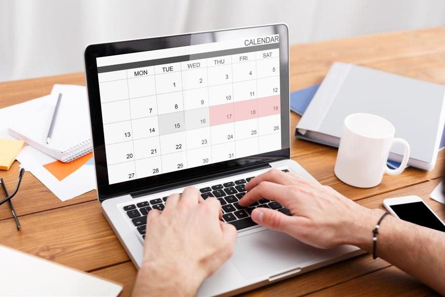 Opened laptop on a wooden desk with a digital calendar showing on the screen and hands typing on its keyboard.