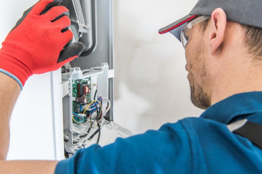 HVAC tech wearing a bright red working glove on his left hand, wearing a blue colored shirt, gray hat, and clear safety glasses, is looking at a furnace's inside wiring.