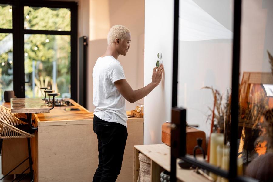 Man wearing a white t-shirt and black pants, adjusting his thermostat that is affixed to a white wall in his house.