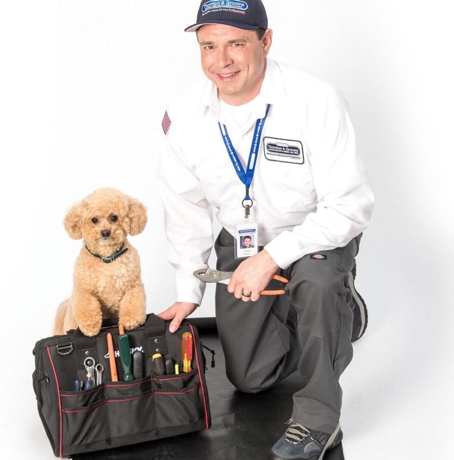 Uniformed Thornton & Grooms tech kneeling next to a tool bag that has a puppy perched on top of it, smiling at the camera, holding a pair of pliers.
