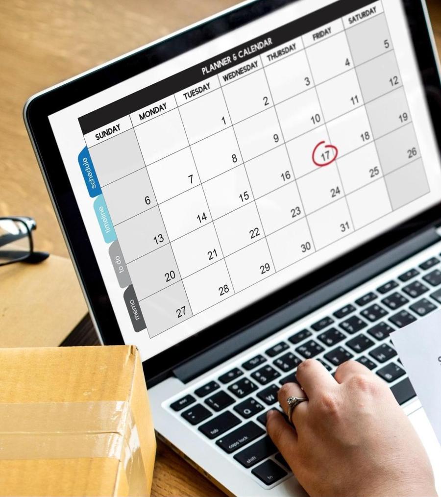 Opened laptop showing a full screen digital calendar on the screen, sitting next to a package and pair of black eyeglasses, with a hand typing on the keyboard.