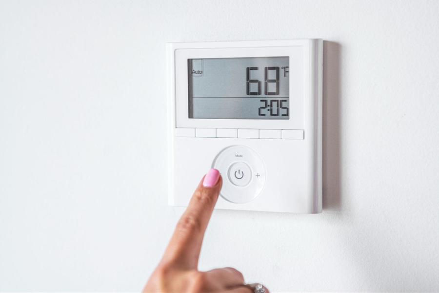 Woman with baby pink nail polish turning her white digital thermostat to 68 degrees, that is affixed to an all white wall.