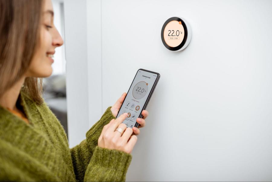 Woman wearing a green sweater, holding her cellphone, smiling at it, adjusting her smart thermostat using an app on her phone and while standing in front of the round digital thermostat on the wall.