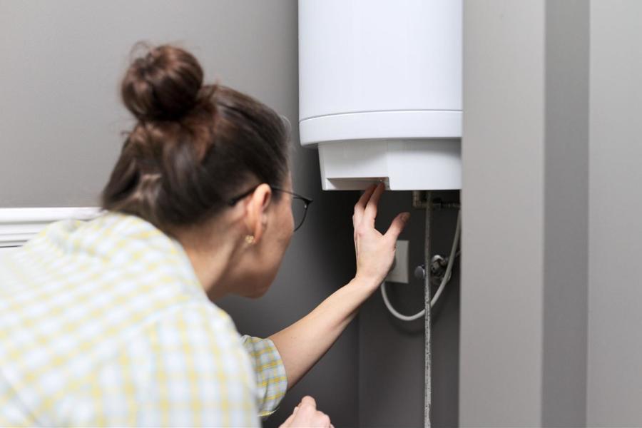 Woman wearing black glasses, a yellow and white plaid shirt, with her brunette hair in a bun, adjusting her water heater’s thermostat