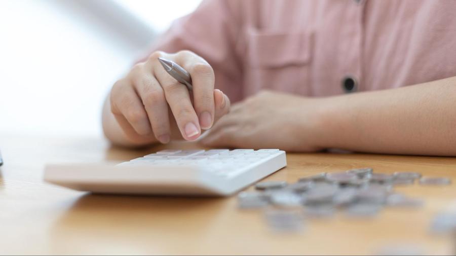 A person holding a pen in their right hand while pressing buttons on a white calculator sitting next to a pile of coins