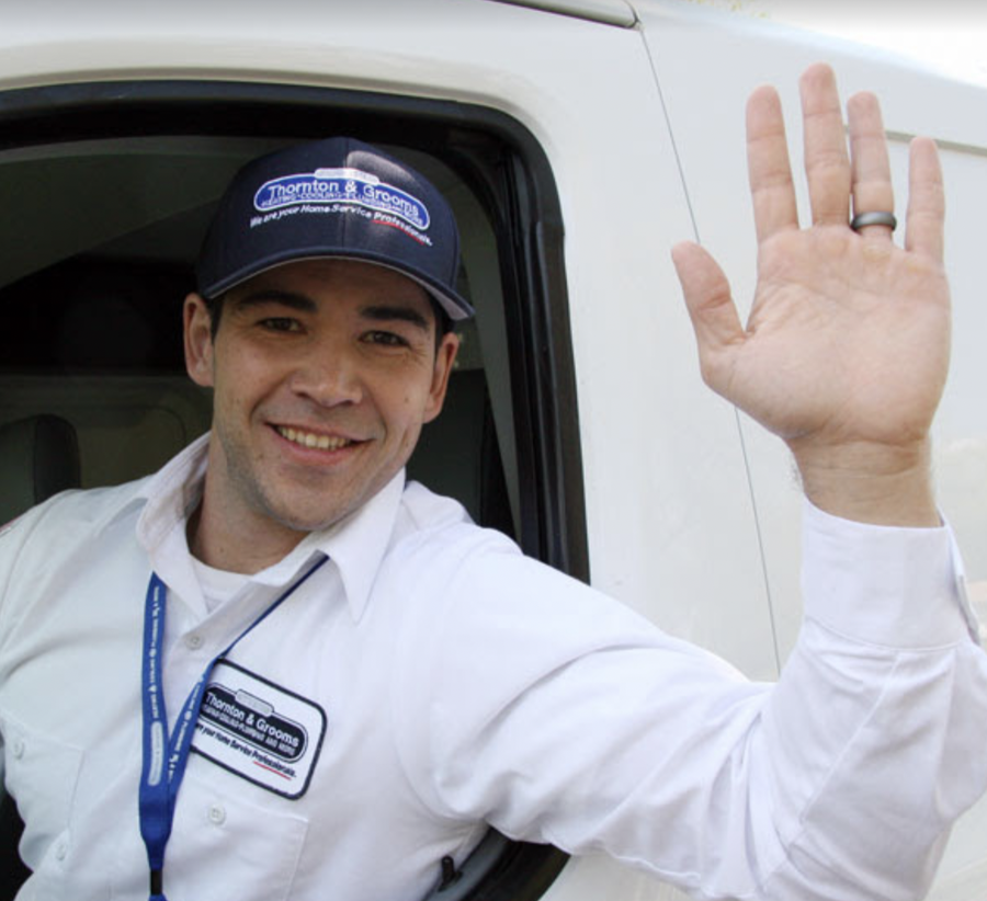 Close up photo of a uniformed Thornton & Grooms tech sticking his left arm out the window and waving and smiling to the camera.