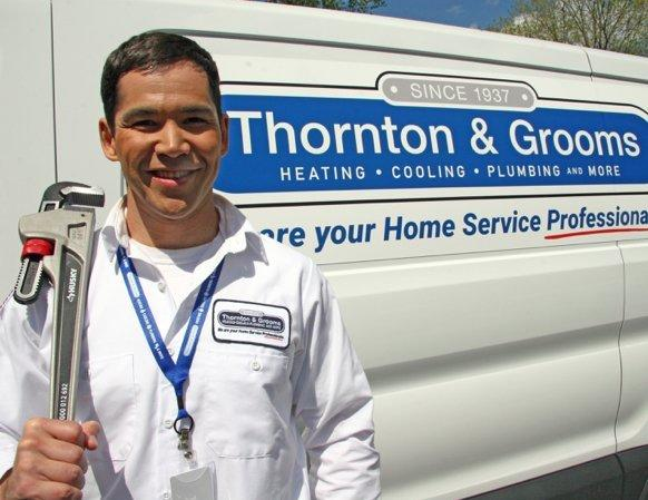 Thornton & Grooms smiling plumber, wearing branded shirt, holding up a pipe wrench with his right hand, standing in front of a branded service van.